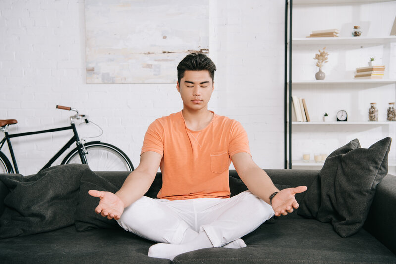 young asian man meditating while sitting in lotus pose on sofa at home
